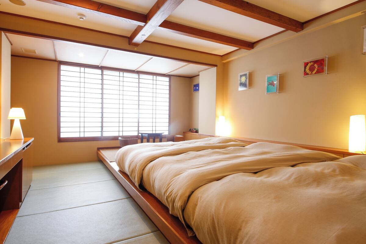 Una habitación japonesa tradicional con suelo de tatami, un futón bajo, un escritorio con lámpara y una ventana con puertas correderas shoji.