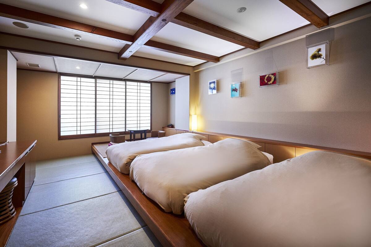 Una habitación tradicional japonesa con suelo de tatami, una cama baja de madera con ropa de cama blanca, una ventana con pantalla corredera shoji y una pequeña mesa con lámpara.