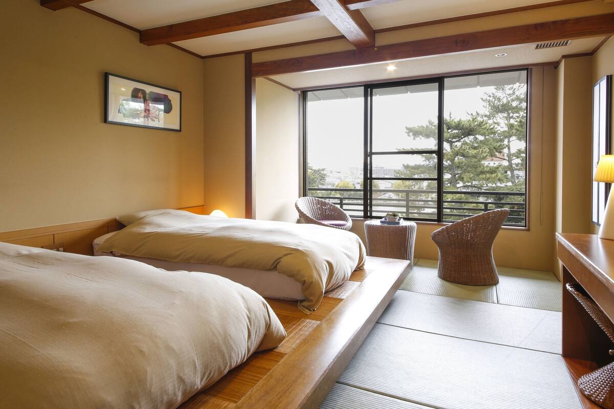 Habitación de hotel con dos camas, un ventanal grande y vistas a árboles.