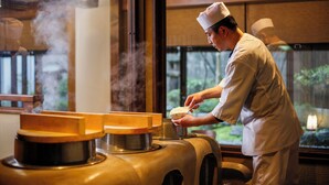 Un chef con uniforme preparando comida en una cocina con una estufa grande y una ventana con vista a un jardín.