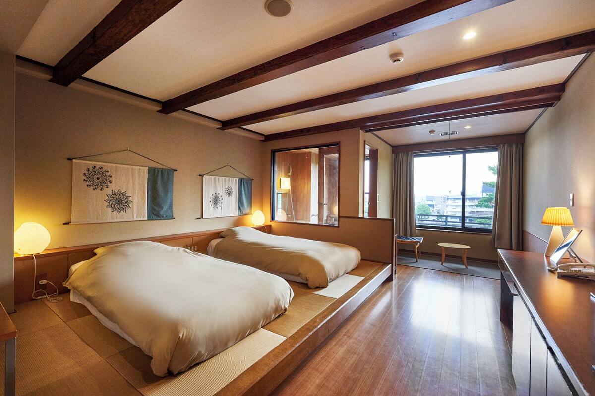 Habitación de hotel con dos camas, una mesita, una lámpara, ventana con vistas y vigas de madera en el techo.