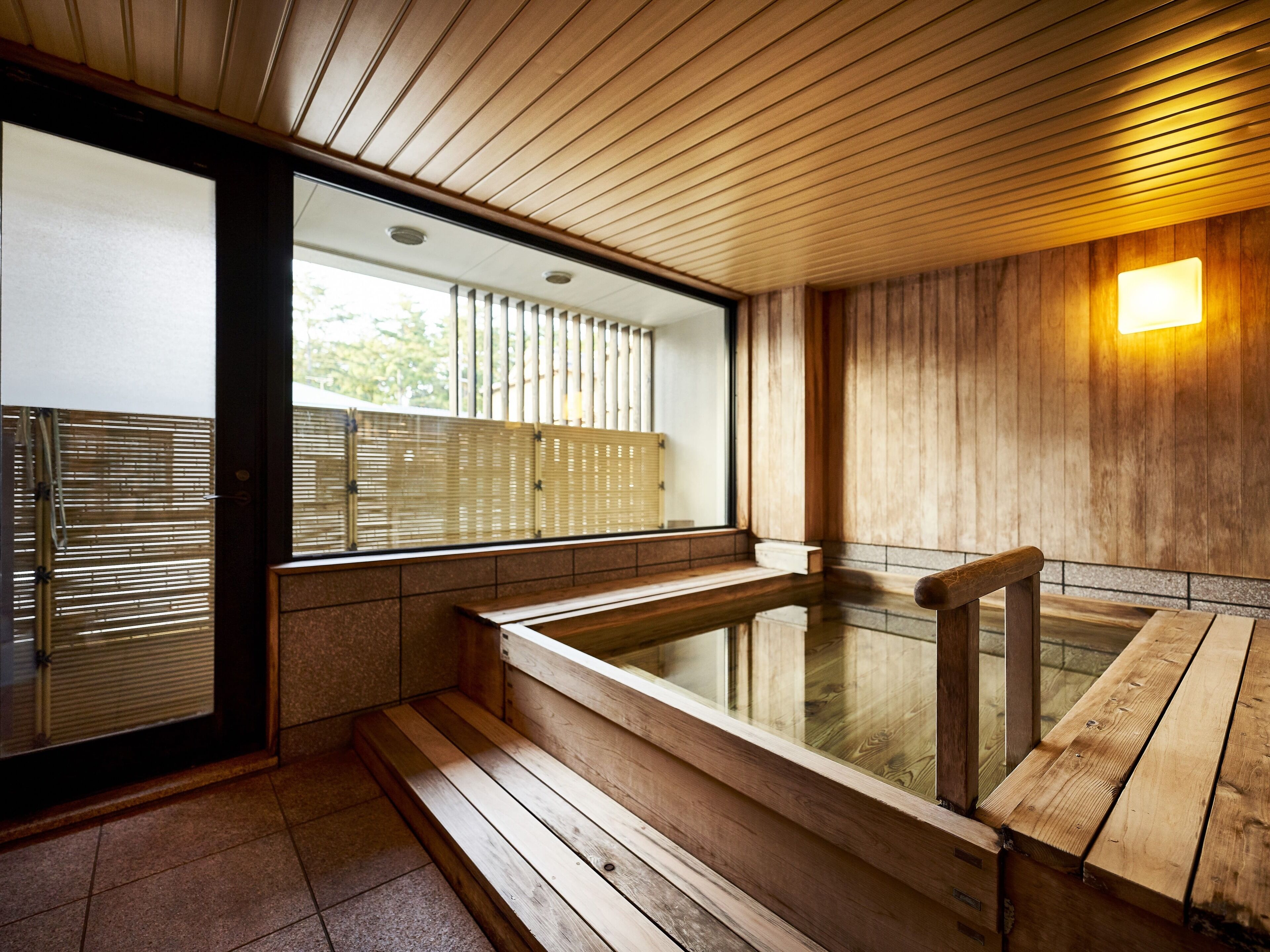 A wooden interior with a hot tub, wooden benches, and a window with blinds.