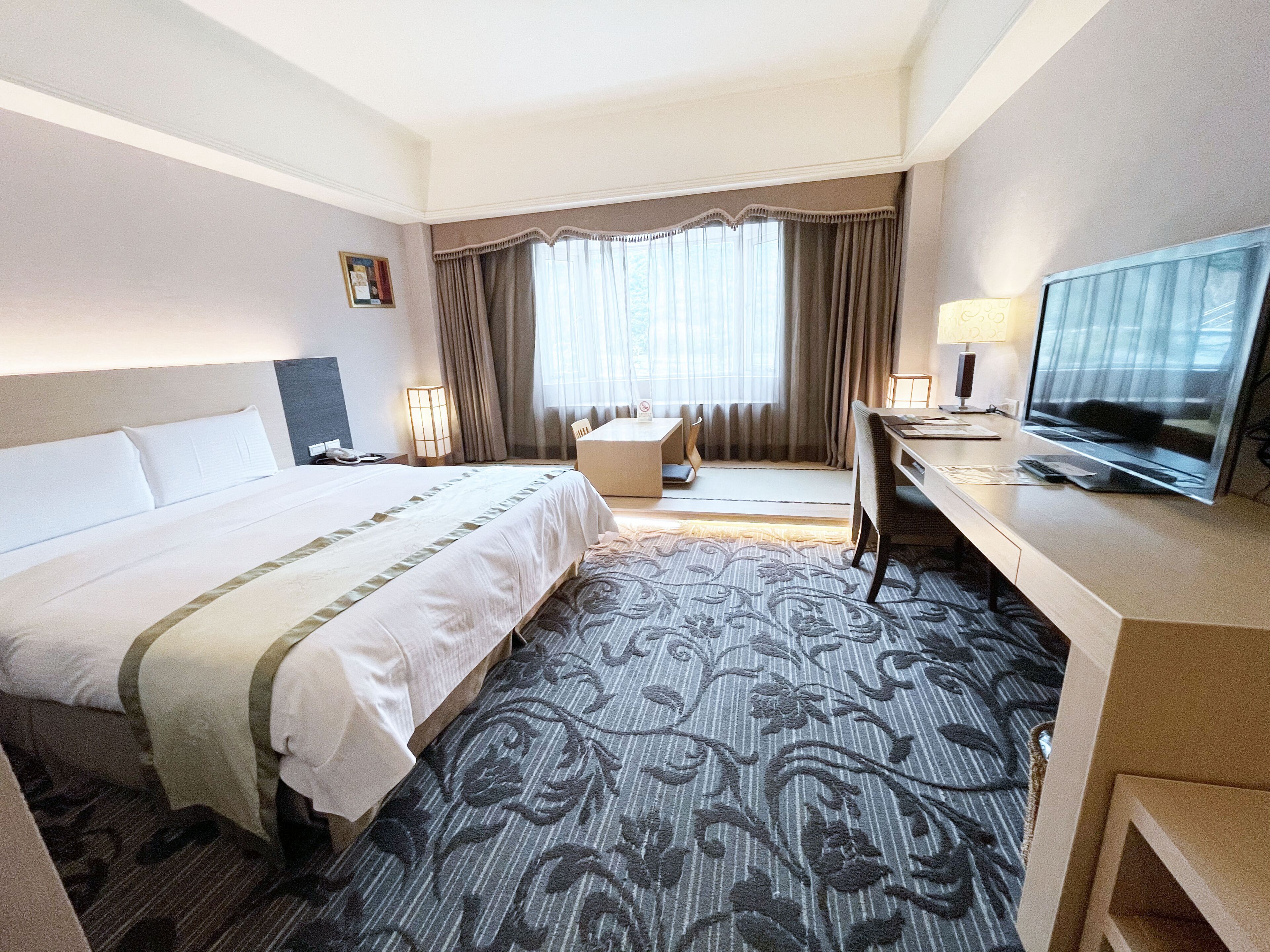 A hotel room with a large bed, a desk with a television, and a window with curtains.