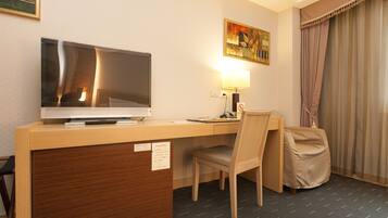 Habitación de hotel con escritorio de madera, televisión, lámpara, silla y cama con cubierta color crema.