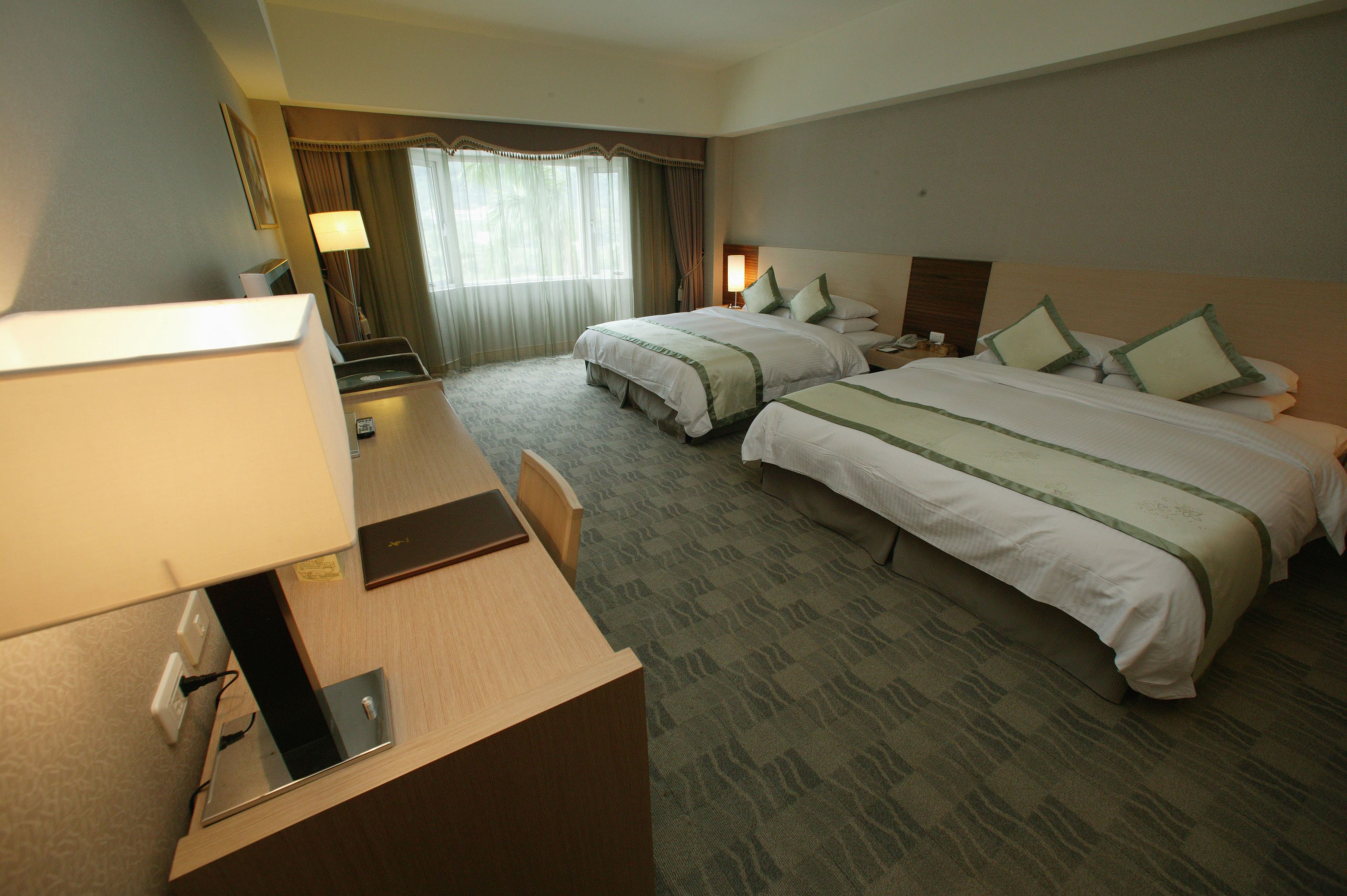 A hotel room with two beds, a desk, a lamp, and a window with curtains.