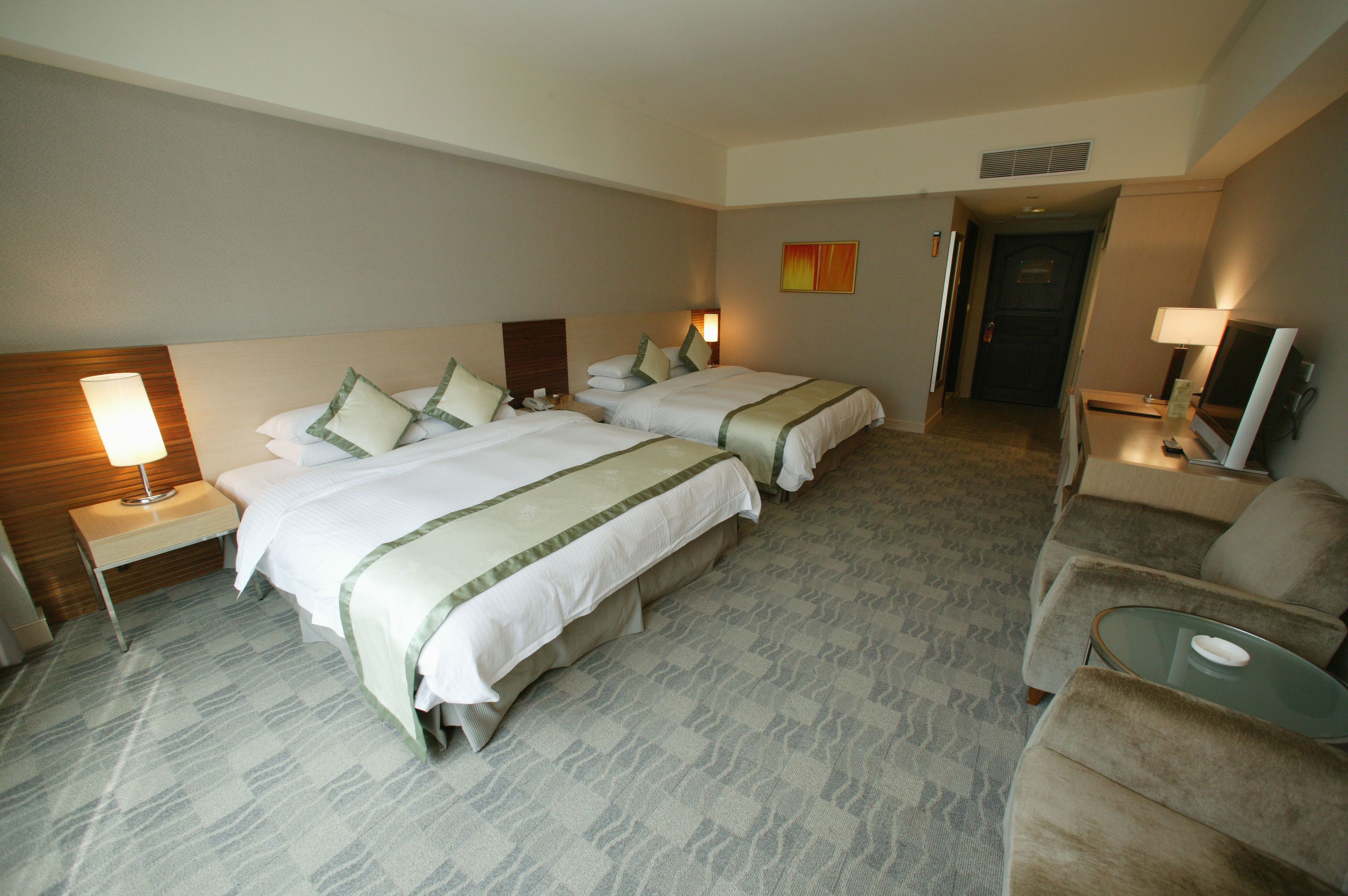 Quarto de hotel com duas camas, um sofá, uma televisão e uma luminária.