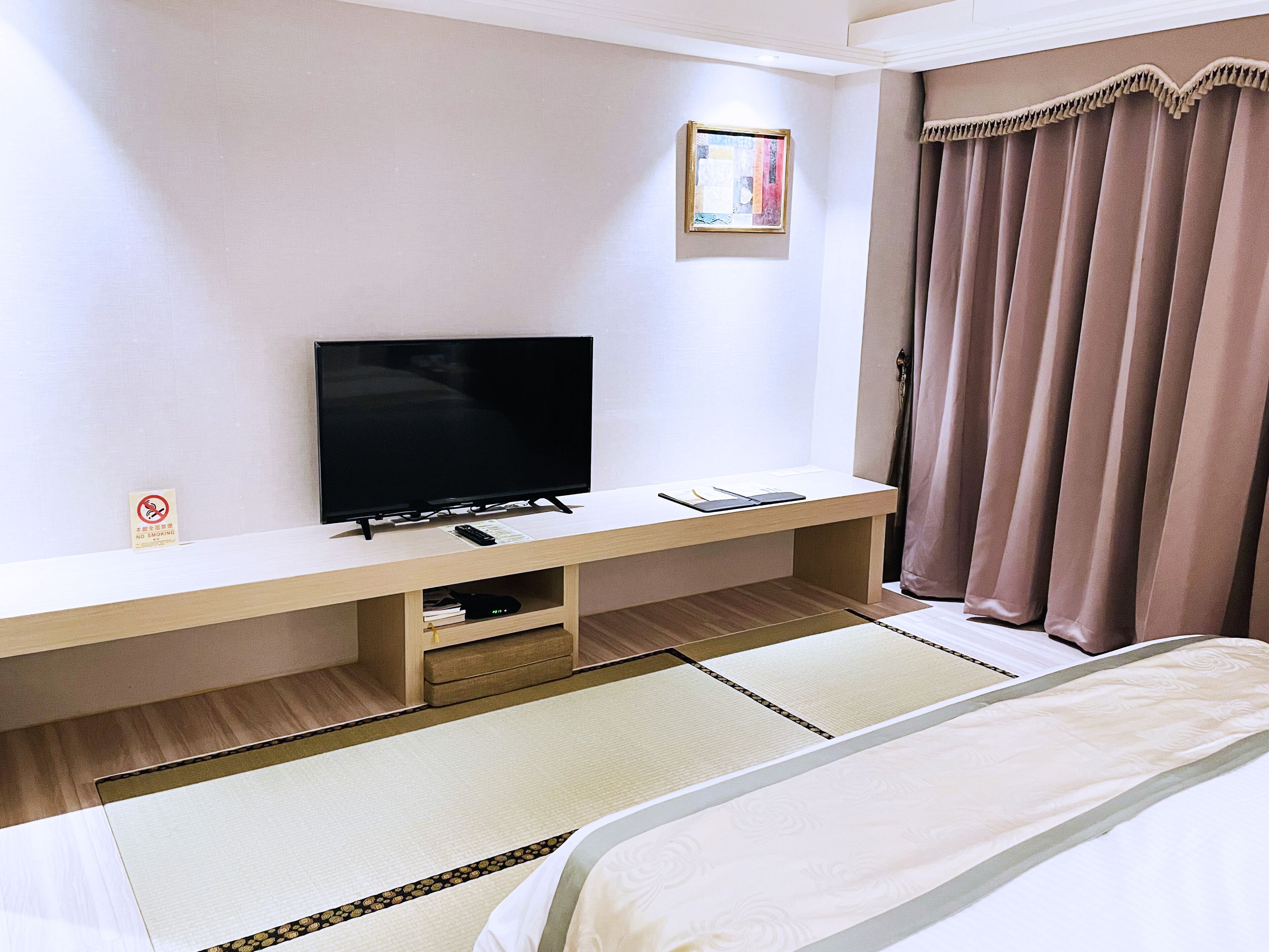 A hotel room with a bed, a TV on a wooden stand, a small shelf with a remote, and a curtain.