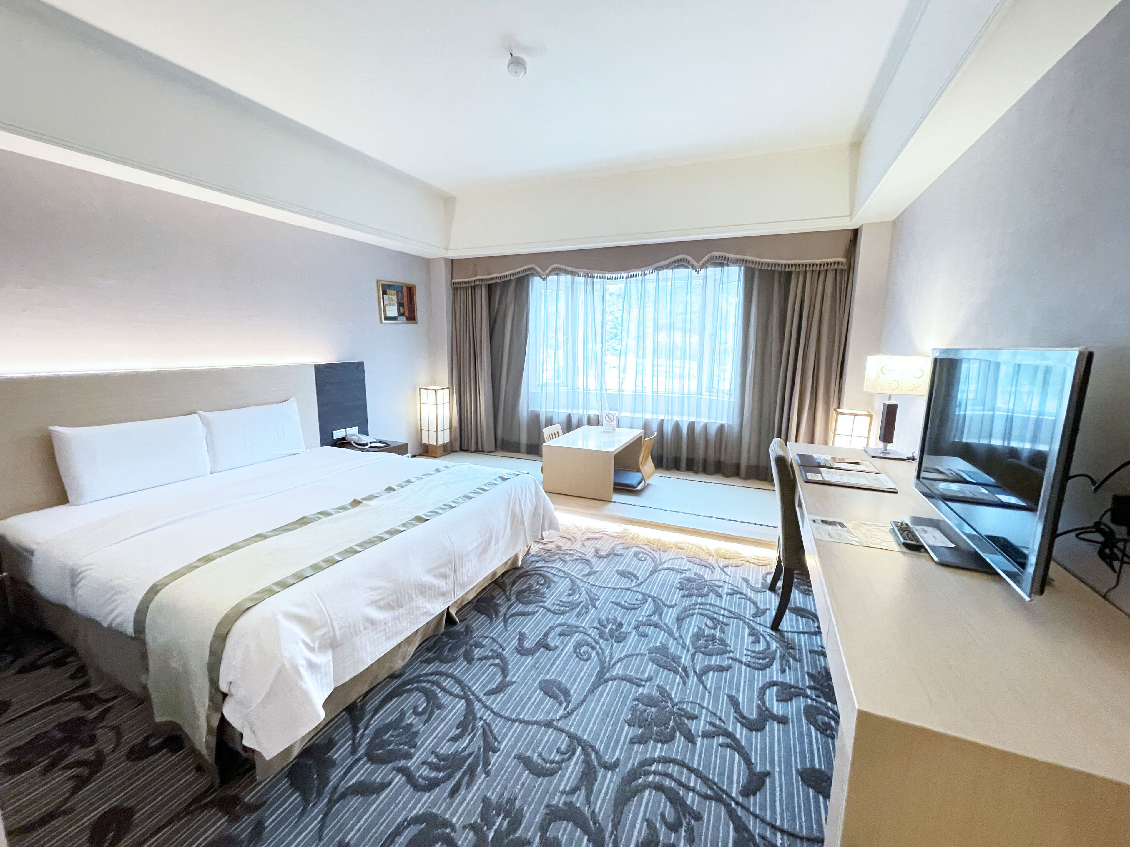A hotel room with a large bed, a desk with a television, and a window with curtains.