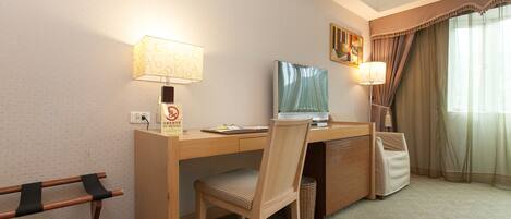 A hotel room with a wooden desk, two lamps, a television, a chair, and a window with curtains.