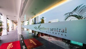 Un área de recepción con un cartel que indica Hotel Boutique Riverfront Sentral, una alfombra roja con el texto 'Welcome' y plantas en macetas.