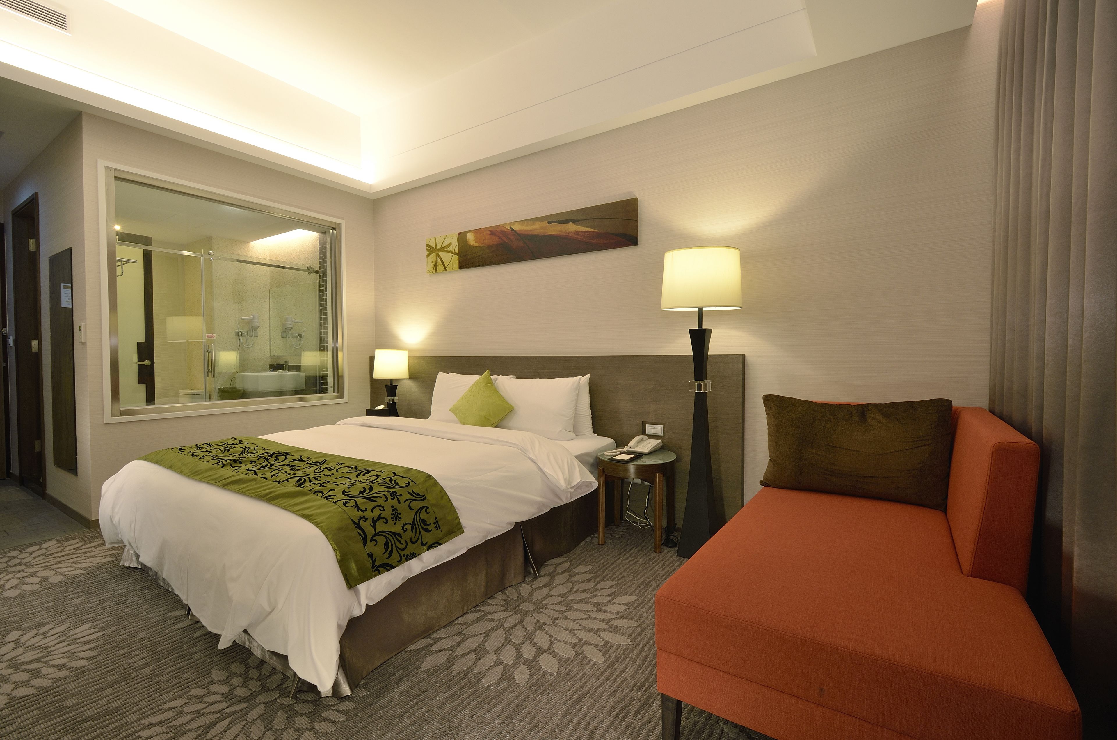 A hotel room with a bed, a nightstand, a lamp, a sofa, and a large mirror.