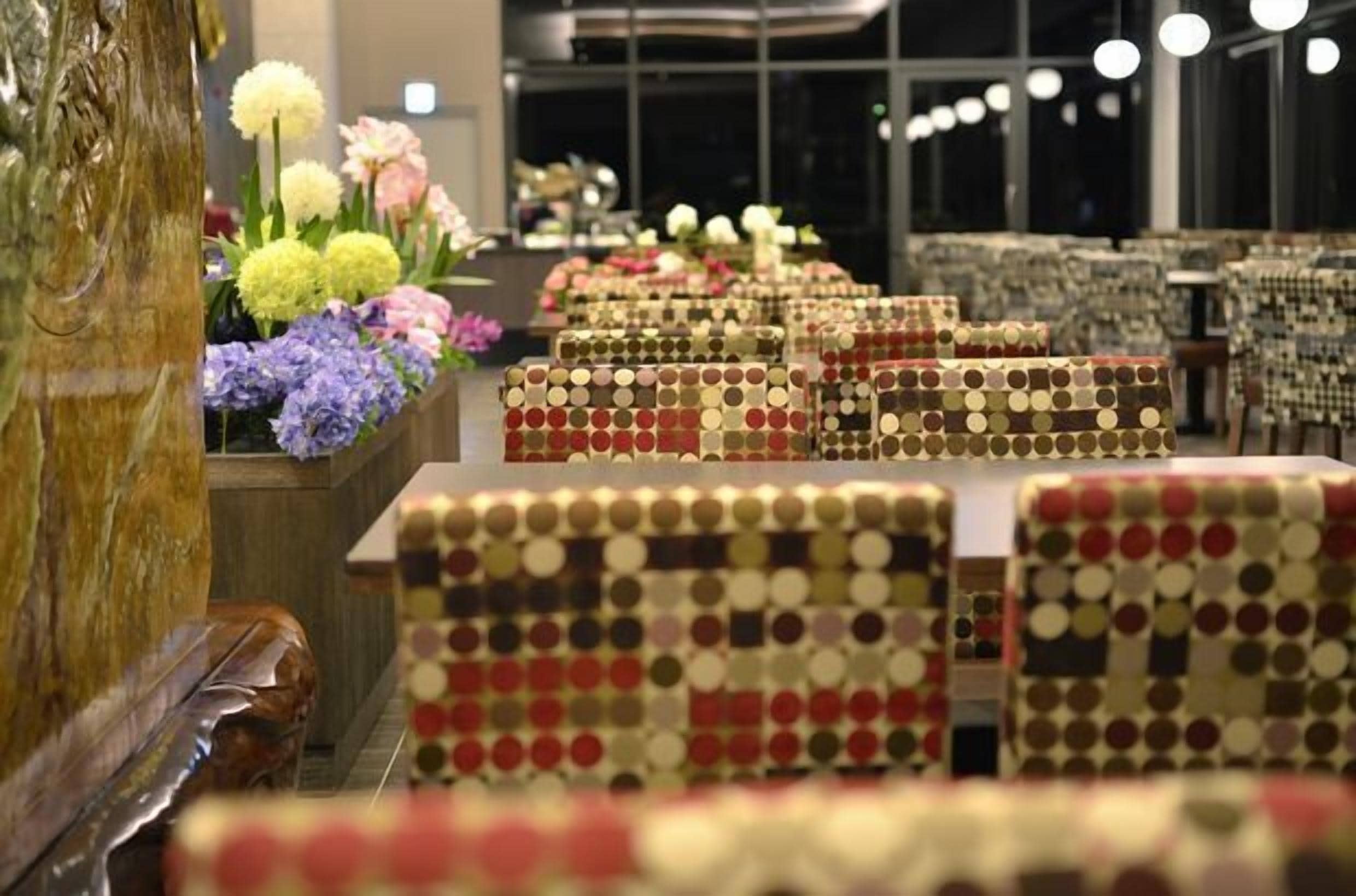 A restaurant with rows of tables covered in patterned tablecloths, a wooden column, and a floral arrangement on the left.
