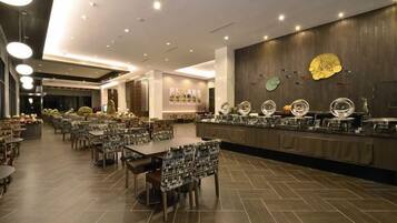 A spacious restaurant with multiple tables and chairs, a long counter with food display, and a modern ceiling design.