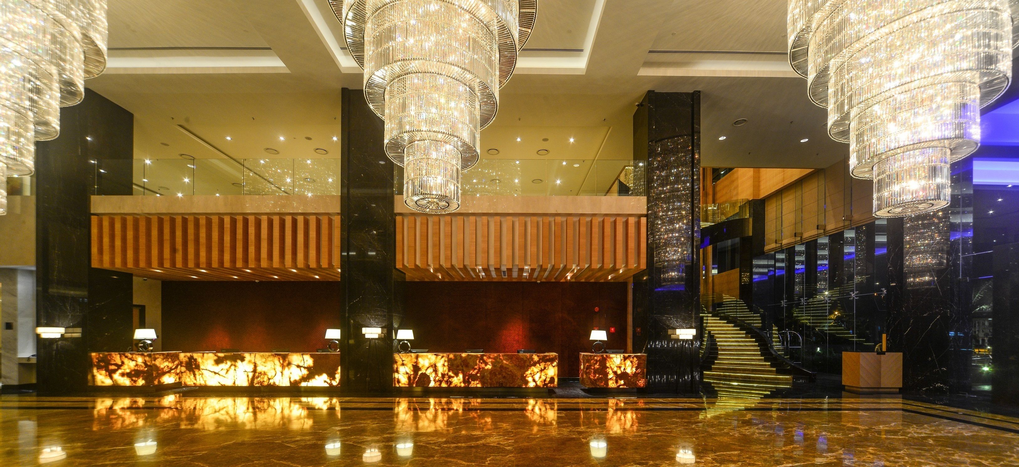 A spacious lobby with a marble reception desk, large chandeliers, and a staircase leading to upper floors.