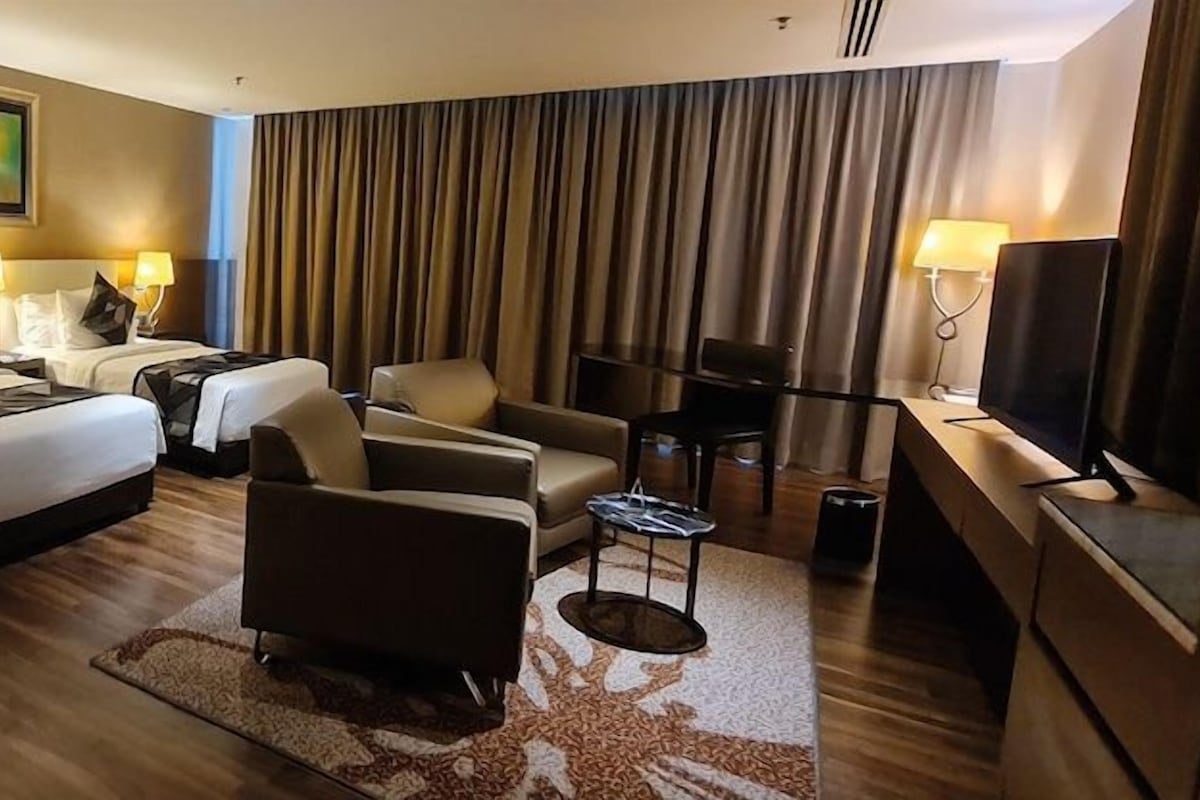 A hotel room with a large bed, two bedside tables with lamps, a sofa, a TV, and a large window with curtains.