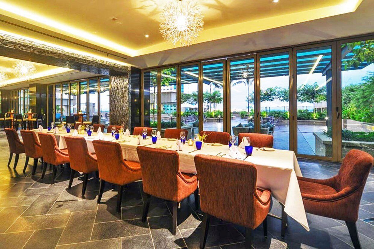 A restaurant with long tables and orange chairs, large windows, and a view of greenery outside.