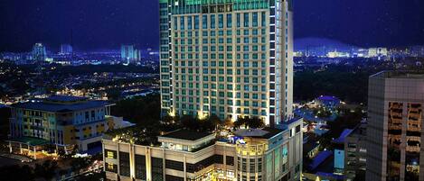 A high-rise hotel with a modern design, featuring a prominent green-lit top section, surrounded by other buildings and a busy urban environment.