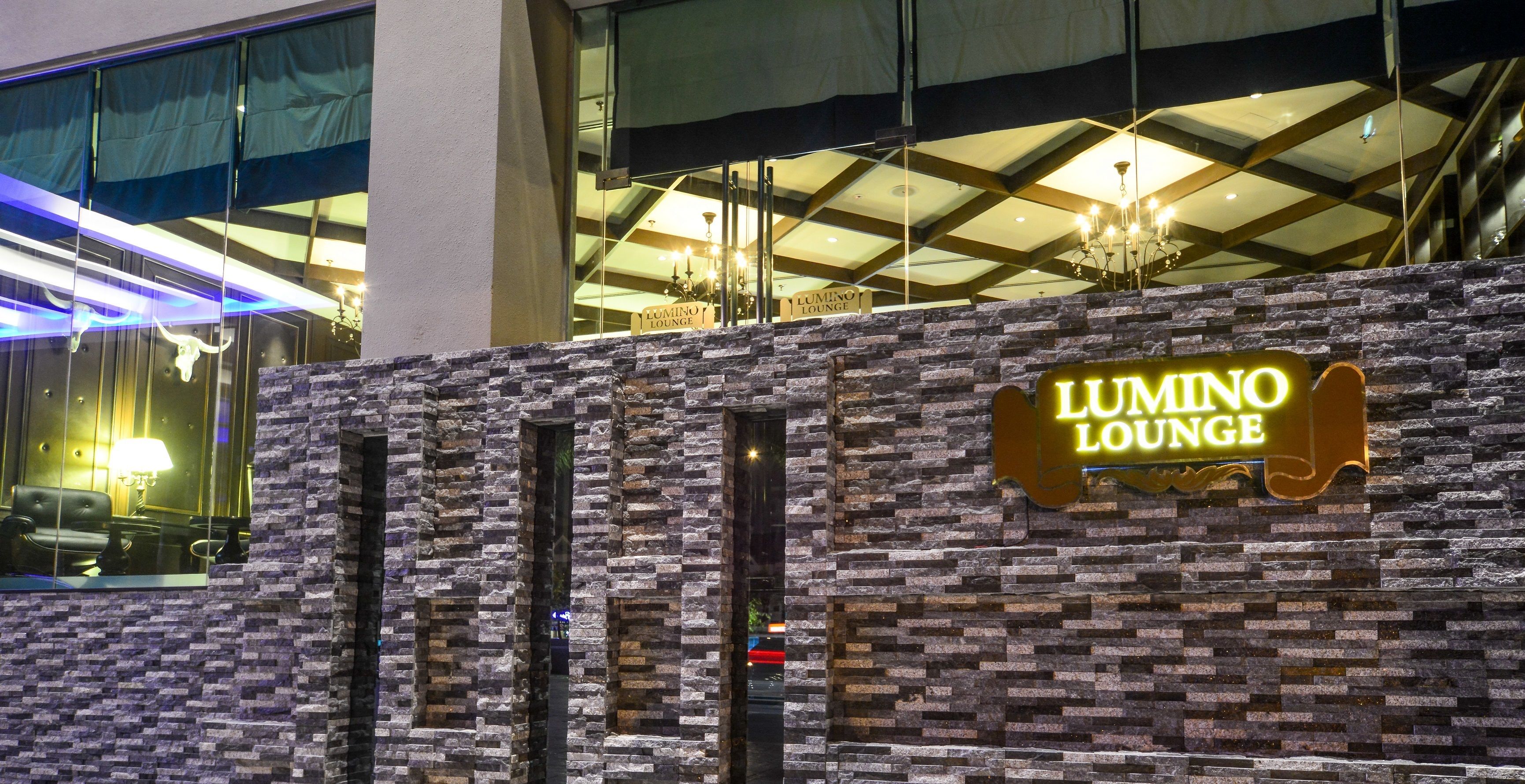A building facade with a stone-textured wall and a neon sign reading "LUMINO LOUNGE".