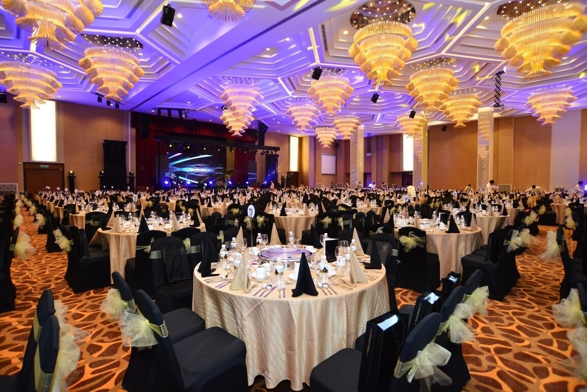 A banquet hall with round tables, black chairs, and chandeliers.