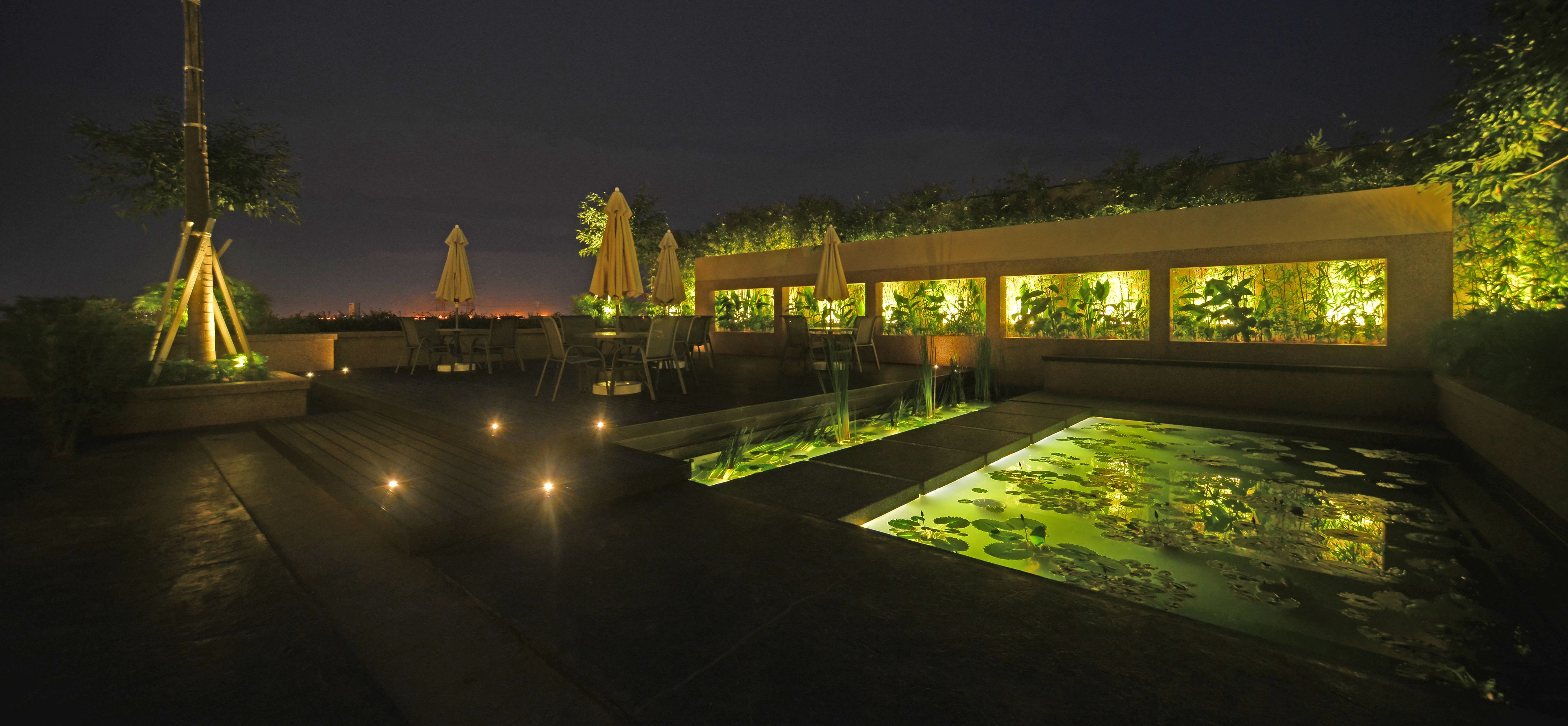 Un espace extérieur aménagé en patio, avec des sièges, un élément décoratif aquatique et des plantes illuminées.