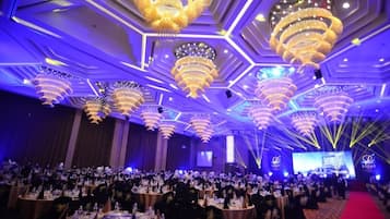 A banquet hall with multiple round tables set for a formal event, featuring a modern ceiling design with geometric patterns and large chandeliers.