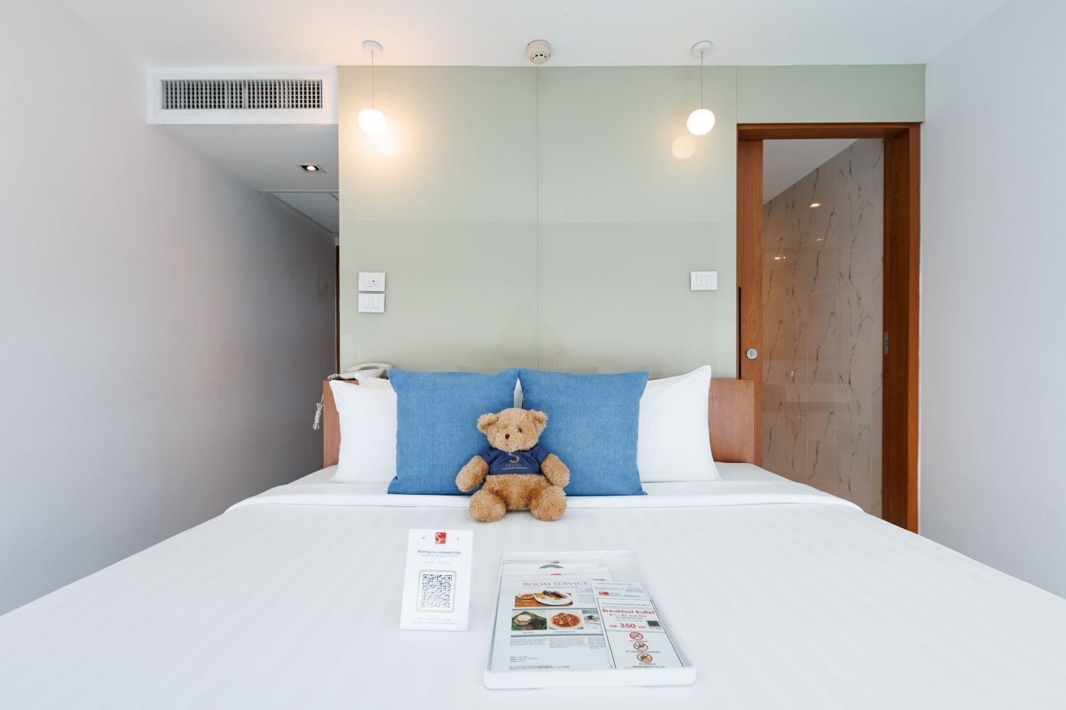 A hotel room with a bed, a teddy bear, a white pillow, a blue pillow, a wooden door, and a wall-mounted air vent.
