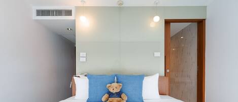 Habitación de hotel con una cama, un oso de peluche, un almohadón blanco, un almohadón azul, una puerta de madera y un respiradero instalado en la pared.