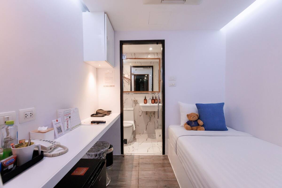 A compact hotel room with a bed, a desk with a telephone and a trash bin, a small bathroom with a sink and mirror, and a teddy bear on the bed.