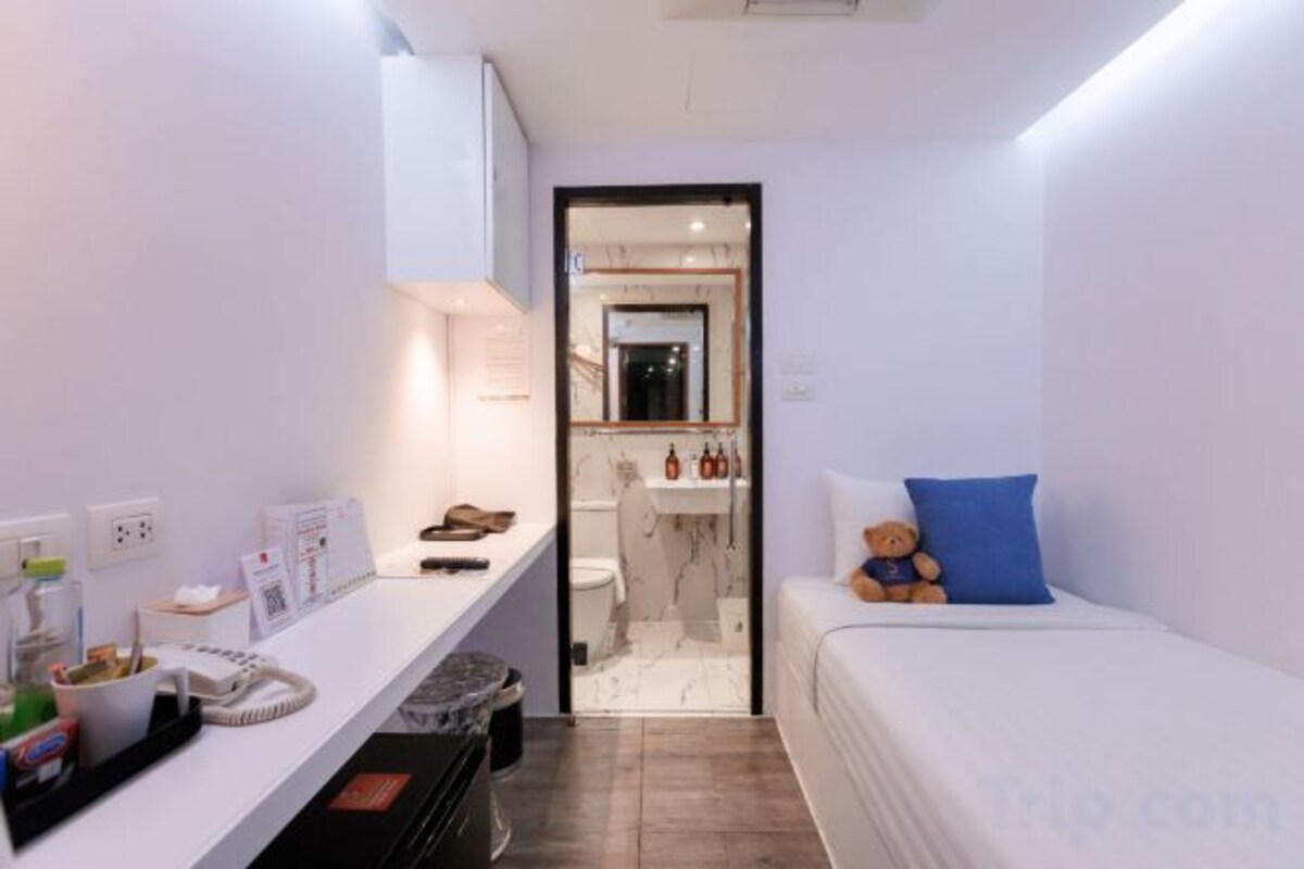 A small, modern bedroom with a single bed, a bedside table, and a nightstand. A bathroom is visible through an open door.