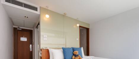 A hotel room with a bed, a teddy bear, a bedside table, a mirror, and a door.