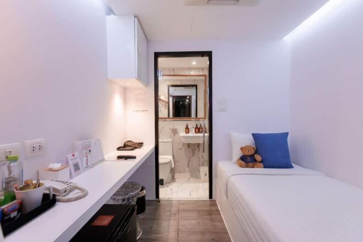 A compact hotel room with a bed, a small desk with a telephone and papers, a mirror, and a bathroom visible through a door.