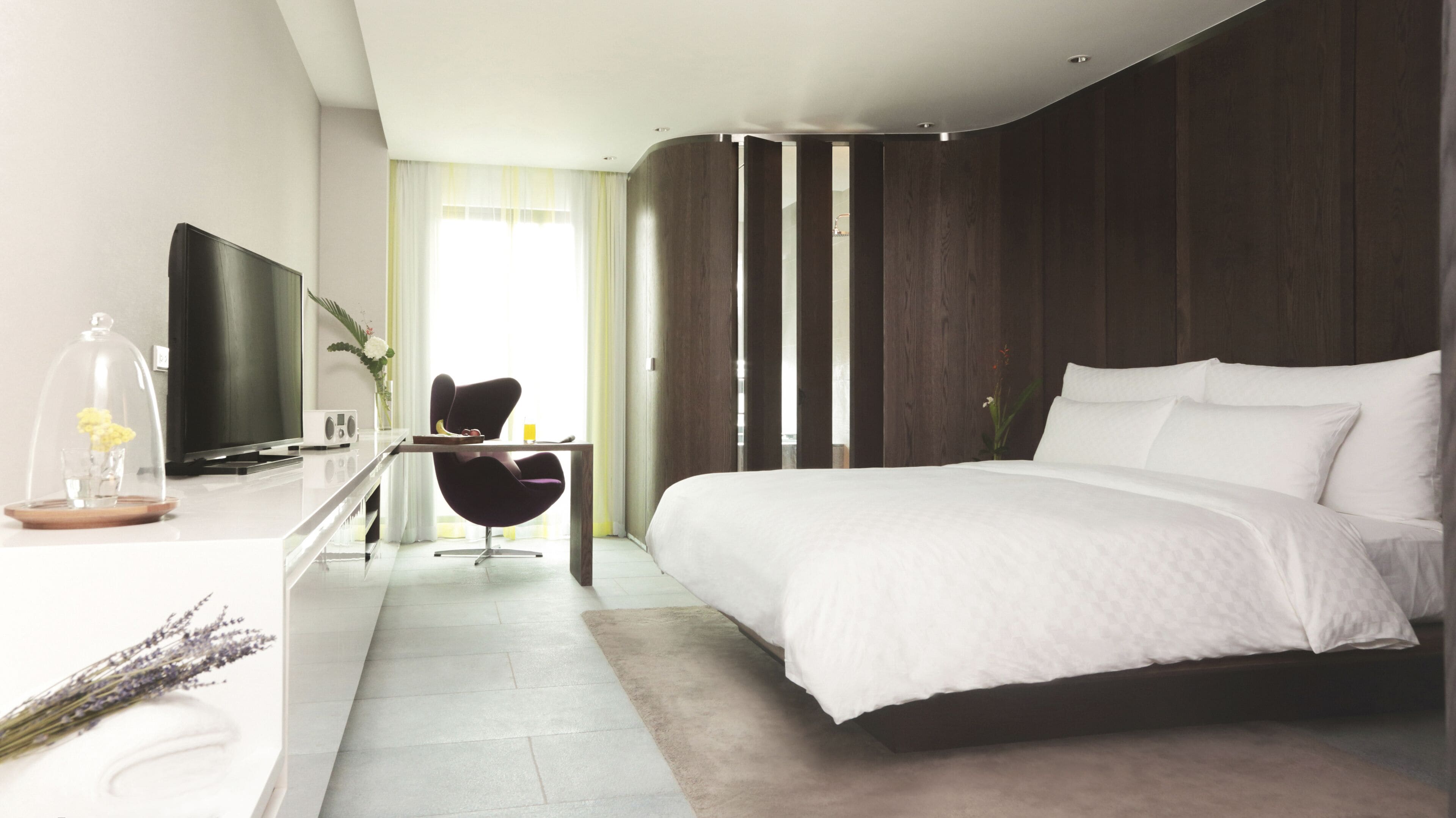 A modern hotel room with a large bed, a desk with a flat-screen TV, a chair, and a view of the outdoors.
