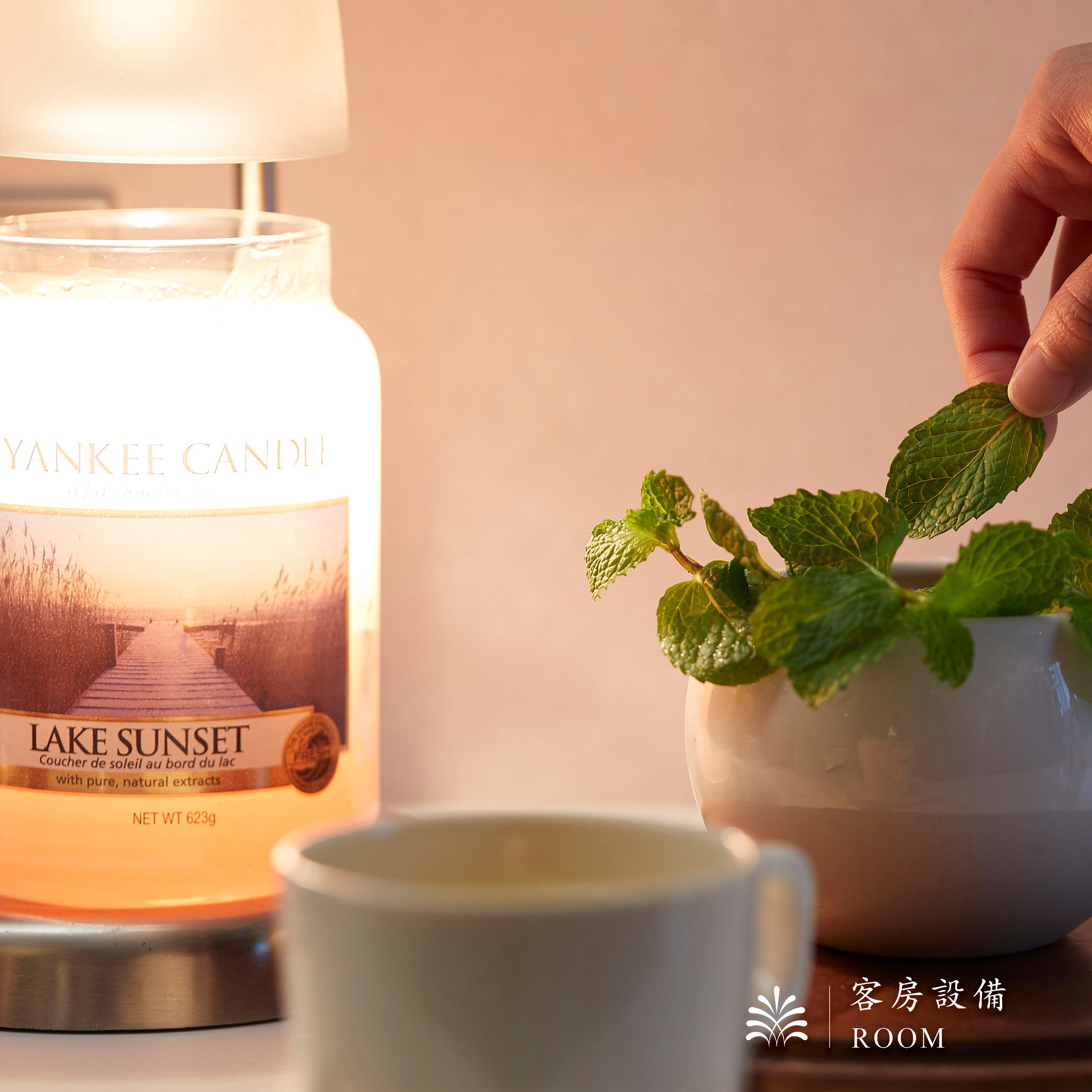 A candle labeled 'Yankee Candle Lake Sunset' with a hand holding a mint plant.