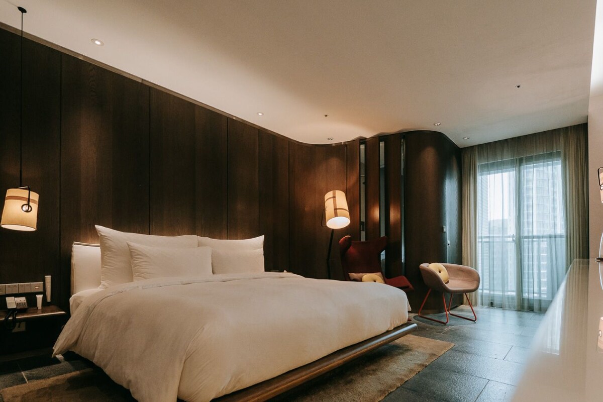 A modern hotel room with a large bed, bedside tables, a chair, and a TV.