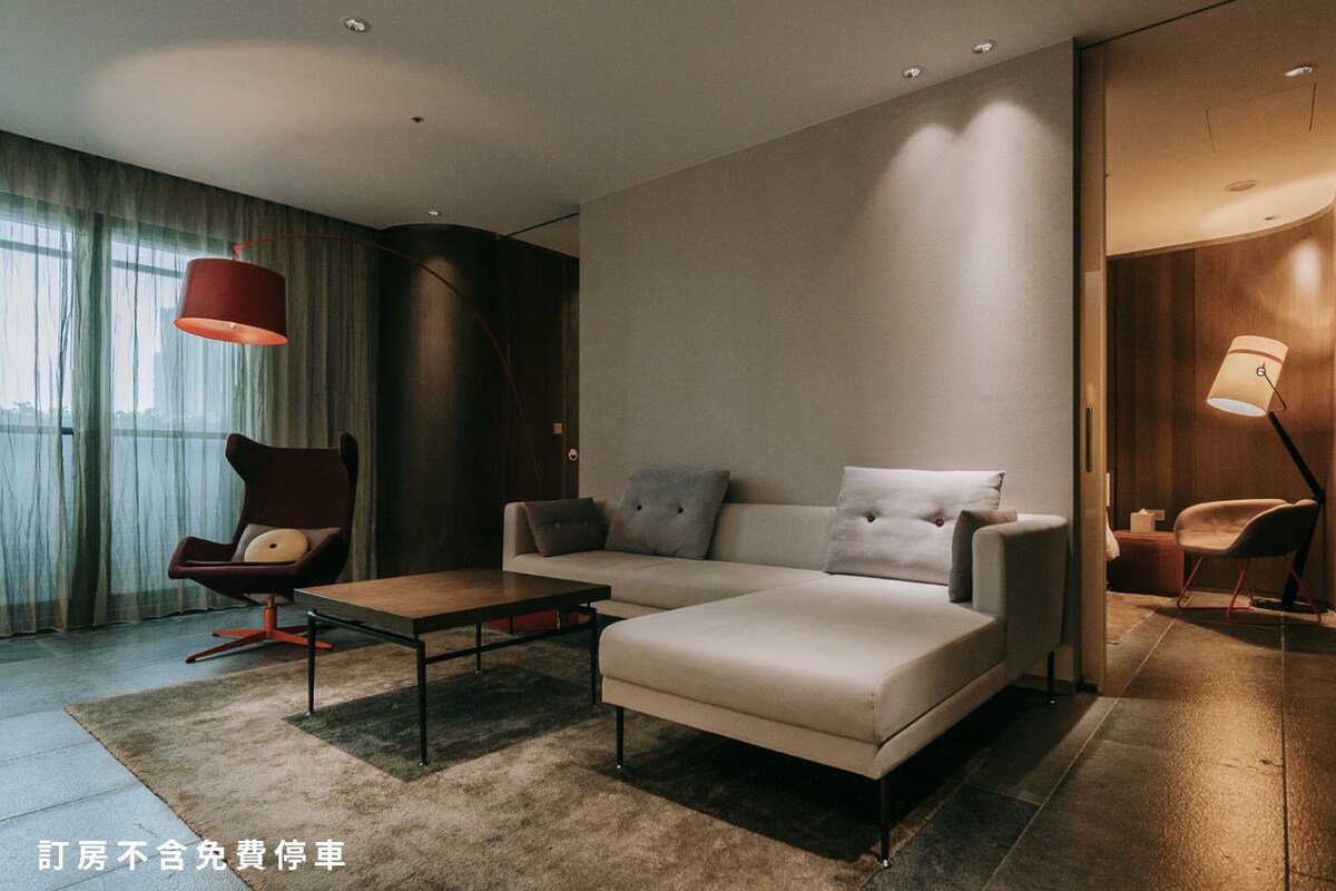 A modern hotel room with a sofa, armchair, and a dining table.