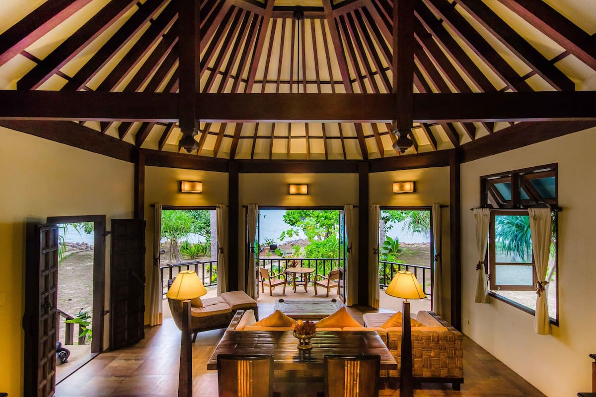 Une pièce spacieuse avec un plafond haut, des poutres en bois, une table centrale entourée de chaises et de grandes fenêtres donnant sur l’extérieur.