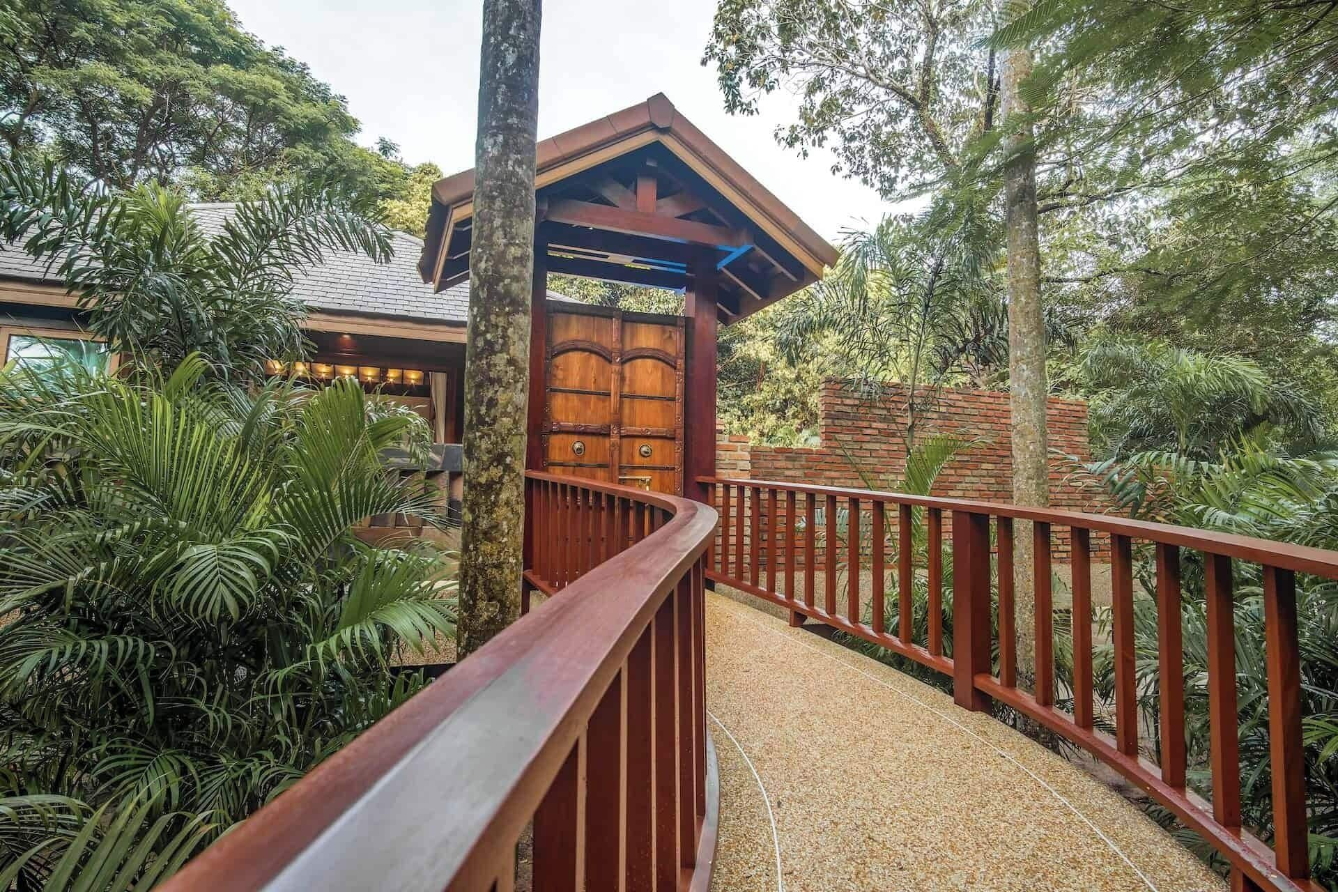 Un sendero de madera que lleva a un edificio con una puerta de madera grande, rodeado de abundante vegetación y árboles.