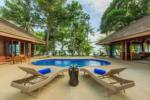 A pool area with lounge chairs, a table with drinks, and a view of the ocean and trees.