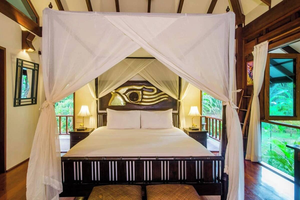 Un lit à baldaquin, avec une tête de lit en bois, des lampes de chevet et une vue sur la verdure grâce à de grandes fenêtres.