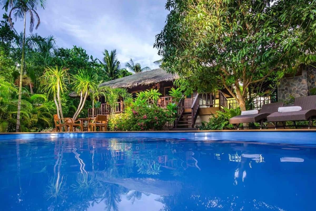 Un espace aménagé au bord de la piscine, avec une structure à toit de chaume, des chaises longues et une végétation tropicale.