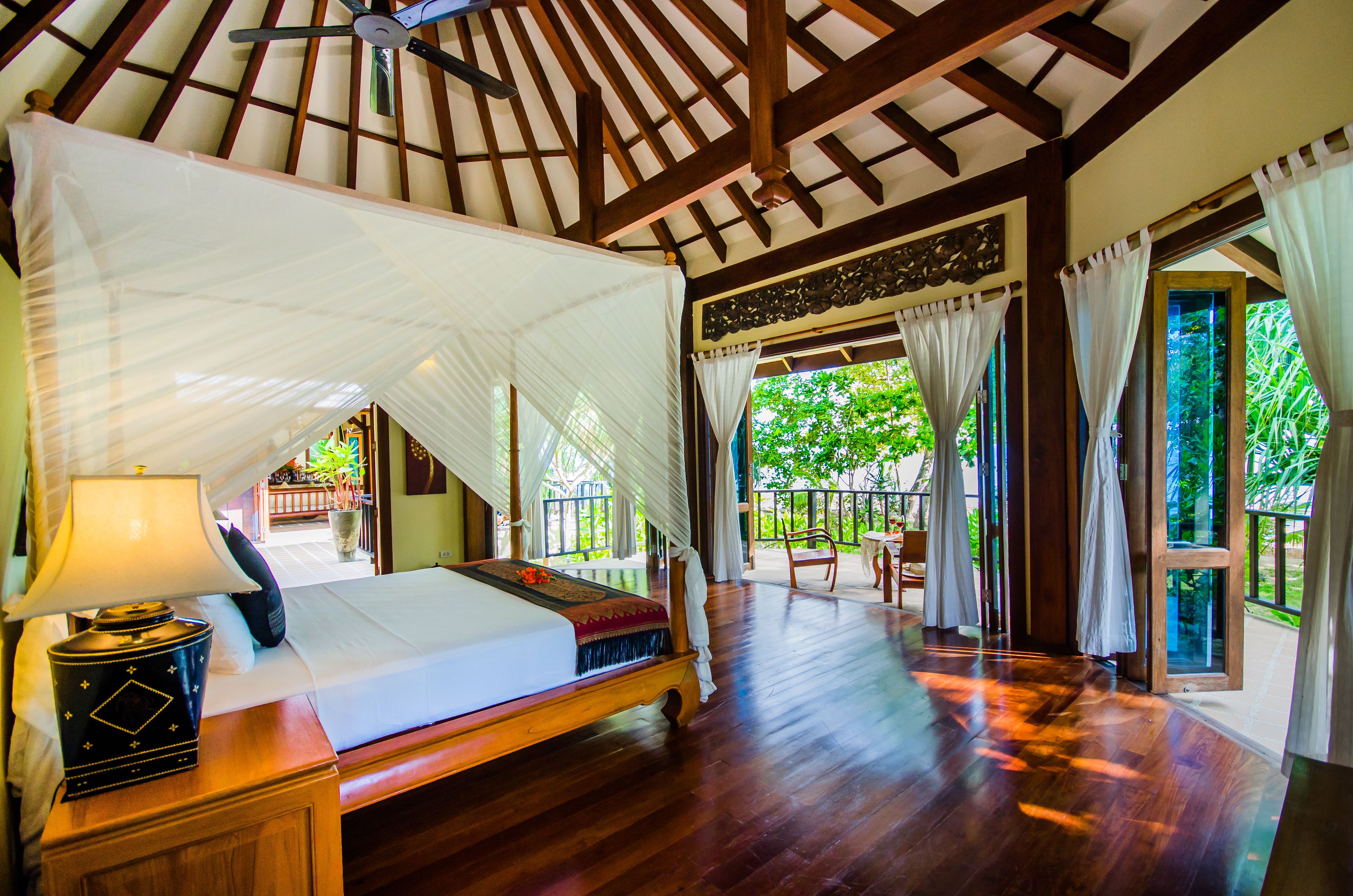 A spacious room with a large bed, wooden flooring, a ceiling fan, and a view of lush greenery outside.
