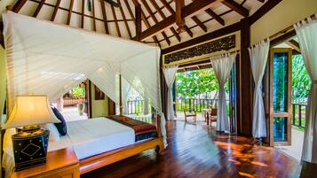 A spacious room with a large bed, wooden flooring, a ceiling fan, and a view of lush greenery outside.