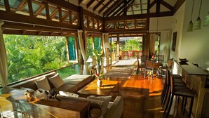 A spacious living area with a wooden floor, a sofa, a dining table, and chairs. There is a view of lush greenery outside through large windows. - Koh Jum Beach Villas “A member of Secret Retreats” (Nuea Khlong)