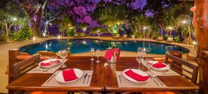 A dining table set for four with a pool view, surrounded by lounge chairs and palm trees.