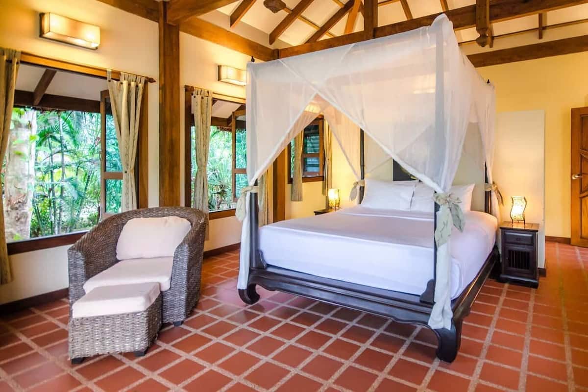 Une chambre à coucher avec un lit sous un dais, une chaise en osier et une table de chevet avec une lampe.