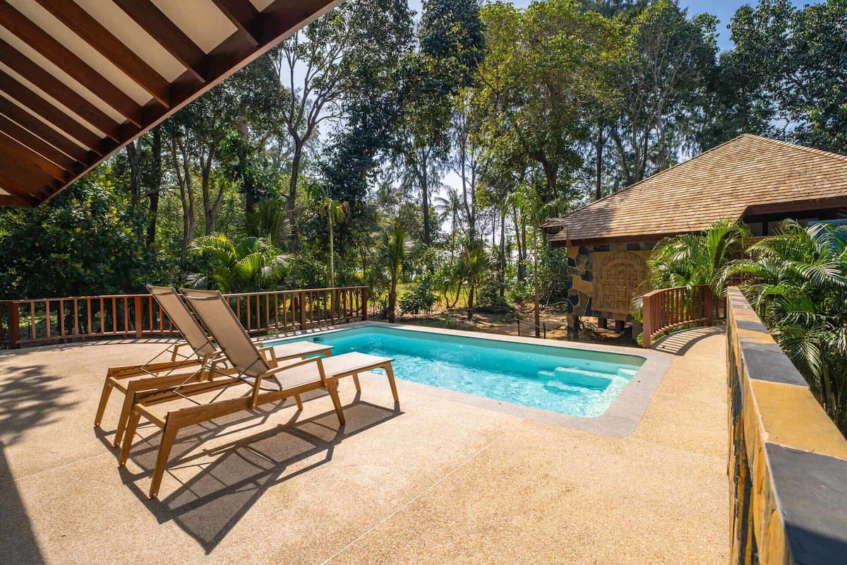 Un espace piscine avec des chaises longues en bois, un bâtiment en pierre et une pergola en bois.