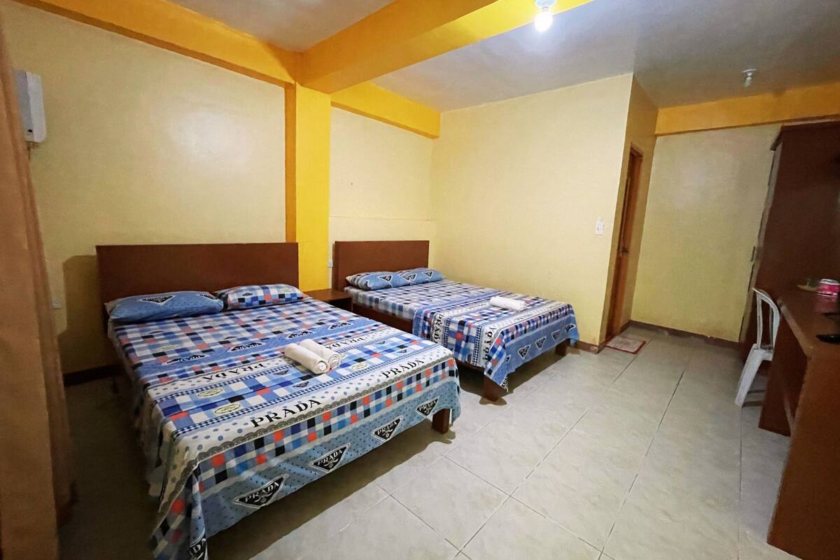 Une chambre avec deux lits, chacun recouvert d’une couverture et doté d’oreillers à carreaux bleus et blancs, avec une tête de lit en bois et un sol carrelé.