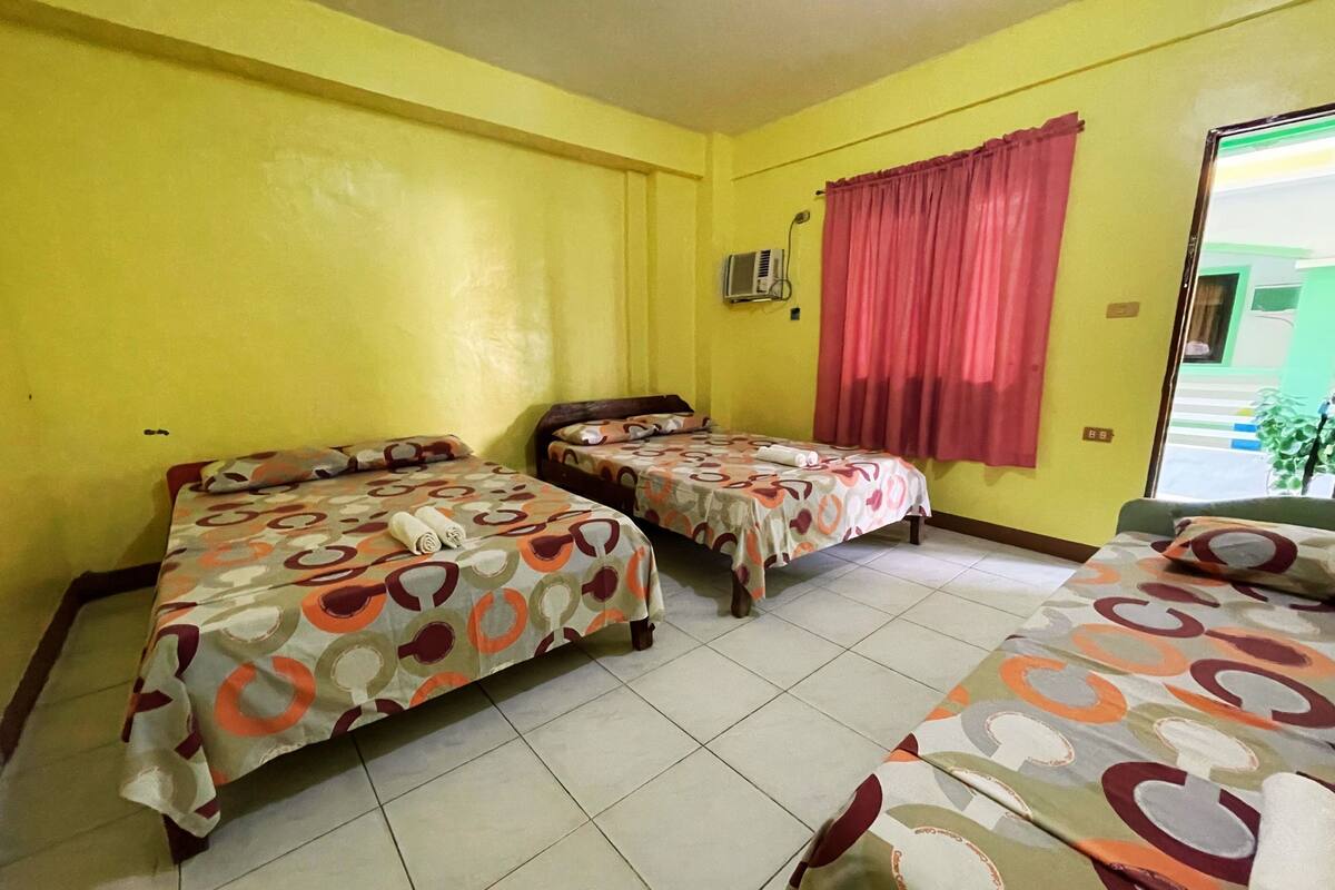 Une chambre avec trois lits, chacun agrémenté de draps à motifs, une fenêtre avec des rideaux rouges et un appareil de climatisation fixé au mur.