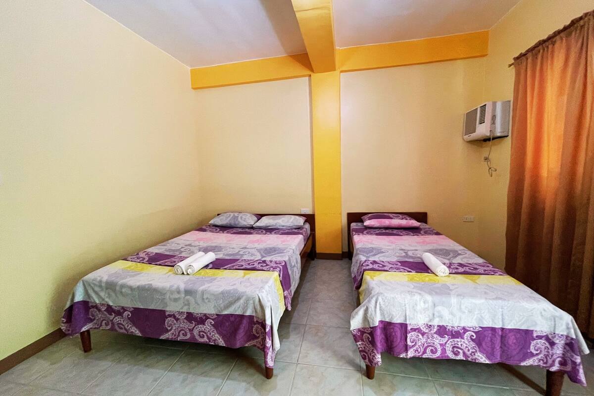 Une chambre dotée de deux lits, chacun recouvert d’une couvre-lit à motifs et agrémenté d’oreillers, ainsi que d’un ventilateur de plafond et d’un appareil de climatisation.