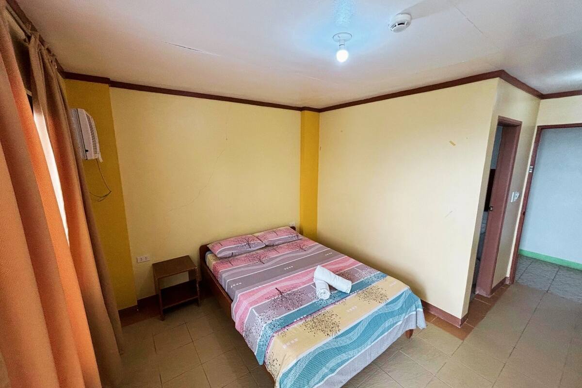 Une petite chambre avec un lit simple, un mur jaune uni, une porte en bois et un sol carrelé.
